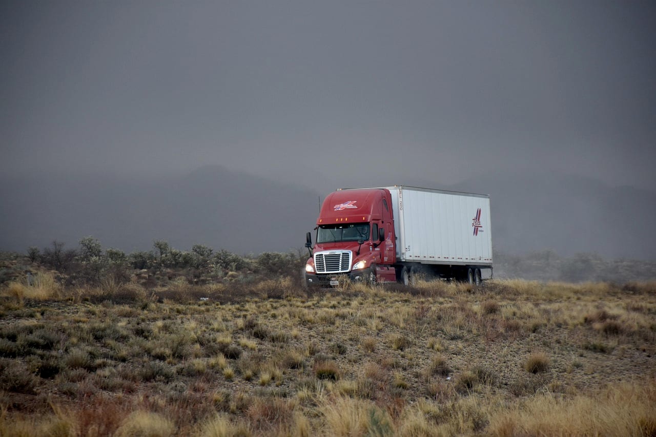 A red tractor-trailer crossing open grassland under a low gray sky